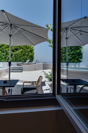 A modern outdoor terrace with white umbrellas, lounge chairs, and a view of a lush green hedge. - Elements Cozy Suites (Santorini)