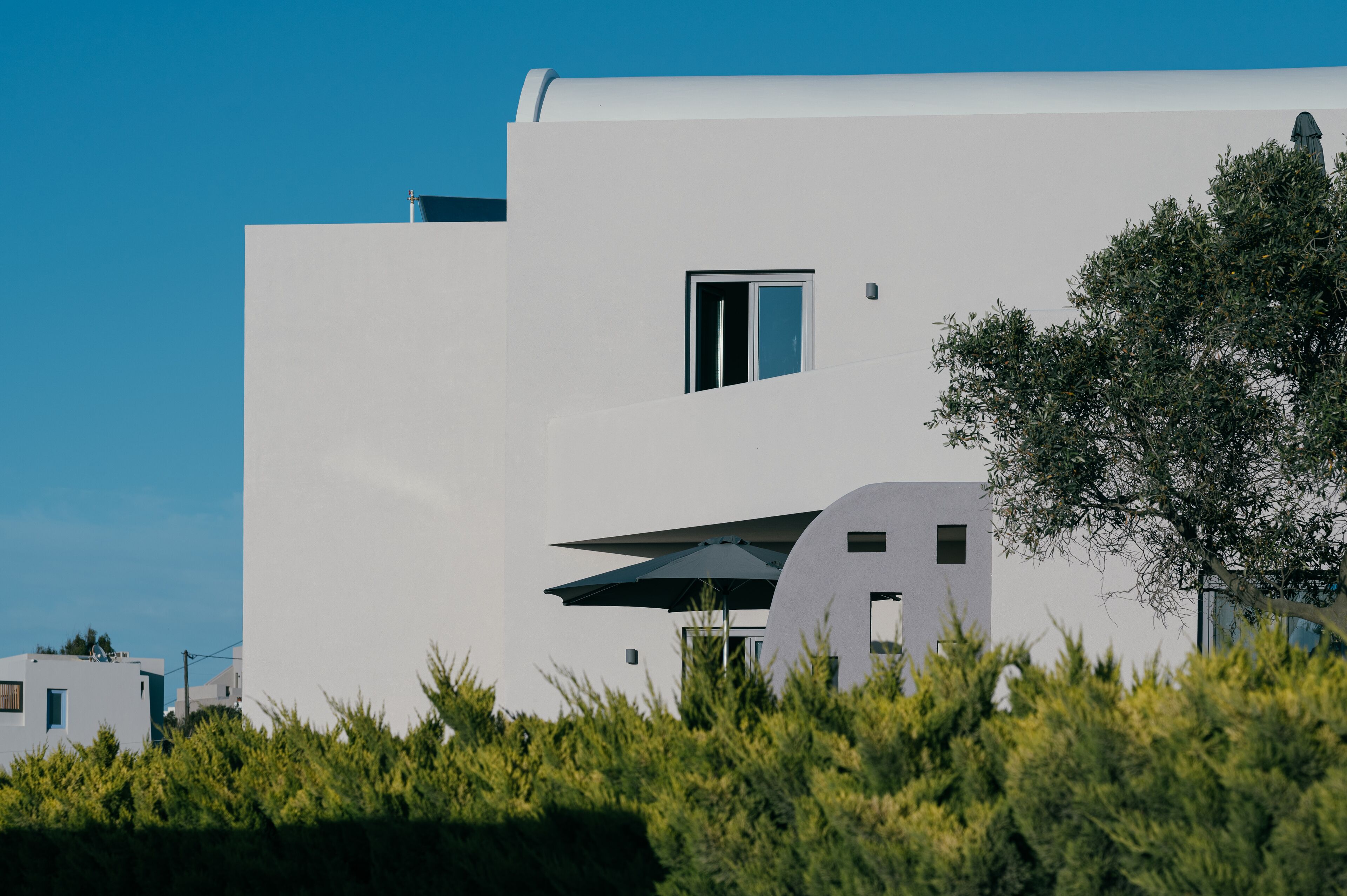 Un edificio moderno blanco con techo plano, una ventana pequeña y un árbol en primer plano.