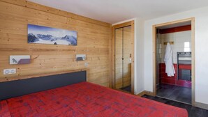 Habitación de hotel con cabecera de madera, cama con colcha estampada roja, pintura de un paisaje montañoso, espejo y zona de ducha con azulejos rojos y blancos.