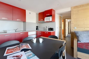 A compact kitchen with red and white cabinets, a microwave, and a coffee maker. There is a dining table with magazines and two white cups.