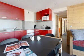 Cocina compacta con armarios rojos y blancos, un microondas y una cafetera. Hay una mesa de comedor con revistas y dos tazas blancas.