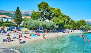 Una playa con agua cristalina, gente tomando el sol y nadando, y árboles al fondo.
