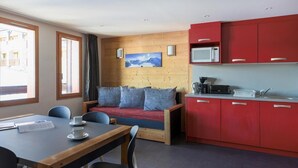 Une pièce de taille modeste avec un mur en bois, un coin cuisine rouge, un canapé avec des coussins, une table à manger avec des chaises et une fenêtre avec des rideaux.