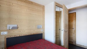 Une chambre à coucher avec une tête de lit en bois, une couvre-lit rouge, une table de chevet avec une lampe et une porte donnant sur une autre pièce.
