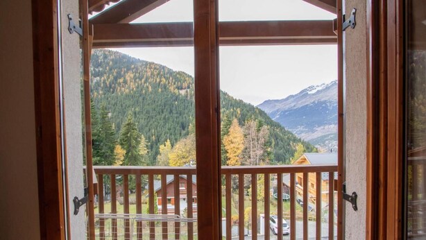 A wooden balcony with a metal railing, overlooking a mountainous landscape with autumn-colored trees and a clear sky.