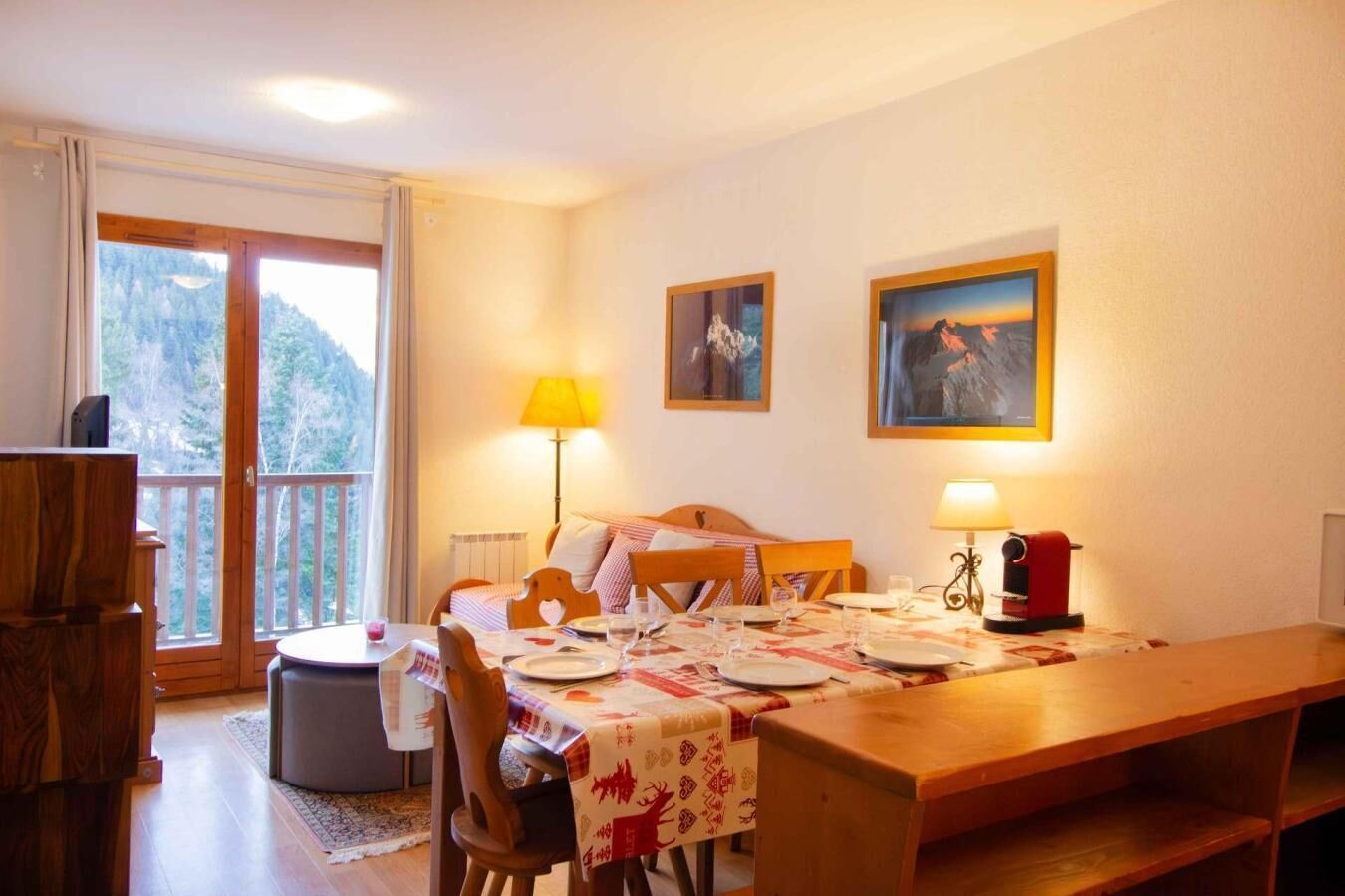 A cozy dining area with a wooden table set for two, a bed with a wooden headboard, and a balcony with a view of trees.