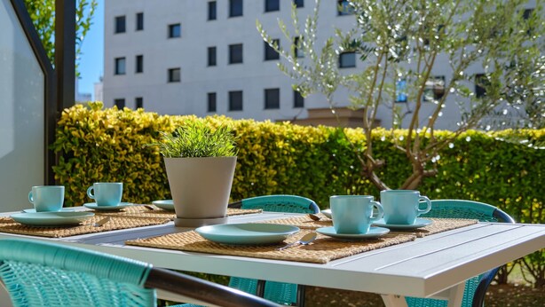 A table set with blue cups and plates, a potted plant, and a view of a building and hedge in the background. - V010 - Milenium, Apartamento con piscina comunitaria y acceso a la playa. (Roses)