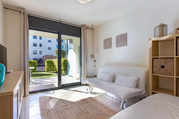 A modern bedroom with a white sofa, a wooden cabinet, and a view of a building through a glass door.