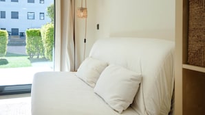 A bedroom with a large window, a bed with white bedding, a hanging light fixture, and a view of a building and greenery outside. - V010 - Milenium, Apartamento con piscina comunitaria y acceso a la playa. (Roses)