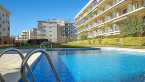 A swimming pool with a metal ladder, surrounded by neatly trimmed hedges and modern apartment buildings in the background. - V010 - Milenium, Apartamento con piscina comunitaria y acceso a la playa. (Roses)
