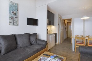 A compact living space with a grey sofa, a small kitchen area, a dining table with chairs, and a flat-screen TV mounted on the wall.