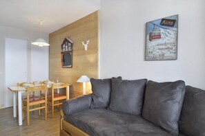 A cozy living room with a grey sofa, wooden dining table, and chairs. A small wooden cabinet with a picture is on the wall, and a lamp is on the table.