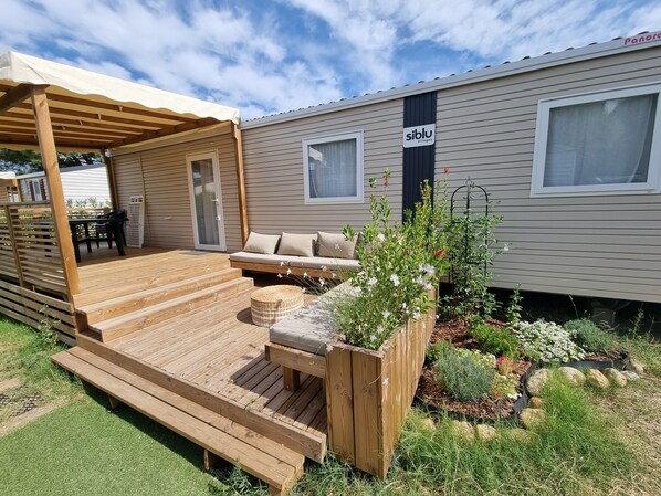 A wooden deck with a sofa and a table, a covered patio area, and a garden with plants and flowers.