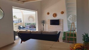 A modern living room with a sofa, a dining table, a TV mounted on the wall, and a fan.
