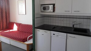 Una cocina compacta con microondas, lavaplatos y fregadero. Un sofá rojo con funda estampada está ubicado junto a la cocina.