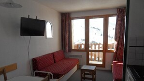 Una habitación compacta con sofá, una mesita, televisor montado en la pared y ventana con vistas a un paisaje nevado.