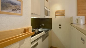 Une cuisine compacte équipée d’une cuisinière, d’un évier et d’un four à micro-ondes. Une étagère en bois se trouve au-dessus de la cuisinière et une bouilloire blanche est posée sur le plan de travail.