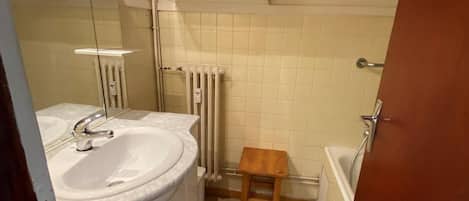 Un baño con lavamanos, espejo, armarios de madera, un radiador y un pequeño taburete de madera.