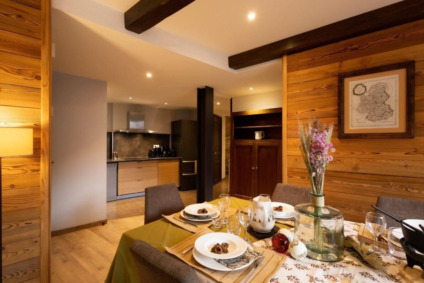 Un coin repas avec une table en bois dressée pour quatre personnes, un vase rempli de fleurs et, au fond, une cuisine.