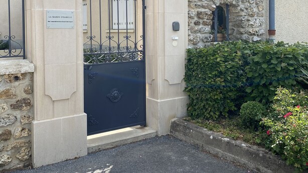 A gated entrance with a stone wall and a metal gate, a stone building with a tiled roof, and a lush green hedge. - Isabelle's Country House (Germaine)