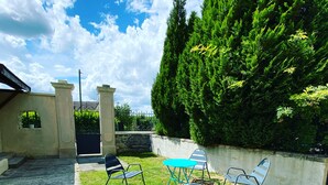 An outdoor patio with a concrete floor, a stone wall, and a metal fence. There are several metal chairs and a small table. - Isabelle's Country House (Germaine)