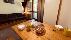 A dining area with a wooden table, bowls, cups, and cutlery. A black sofa and a TV are visible in the background. - 250m from ski slopes, 3rd floor, balcony, tv, ski locker, 20m², Peisey-Vallandry (Landry)