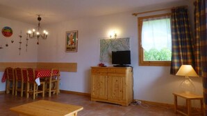 A cozy living room with a wooden dining table, chairs, a TV on a wooden cabinet, a map on the wall, and a window with curtains.