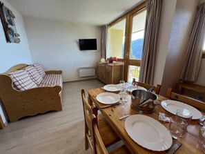A dining area with a wooden table set for four, a wooden sofa, and a television mounted on the wall.