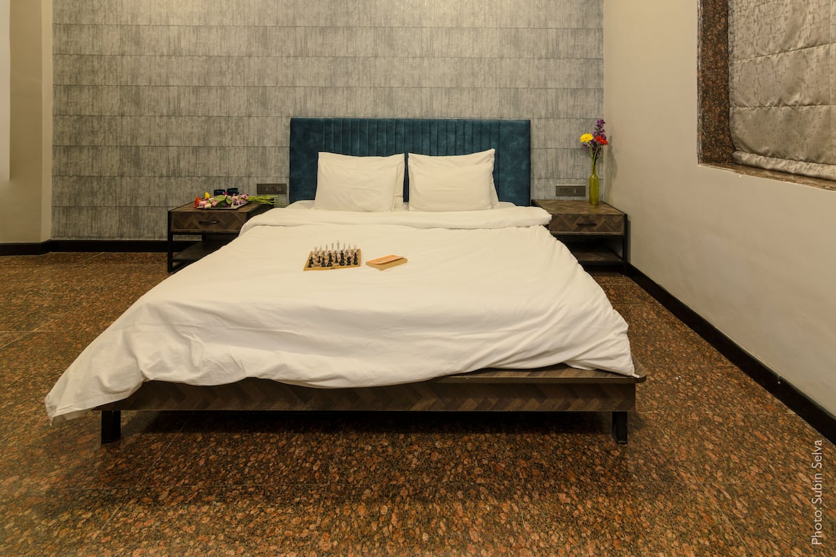 A neatly made bed with white linens, a blue headboard, and a chessboard on the bed. There is a nightstand with a vase of flowers and a clock. The room has a textured wall and a window with a view of the outside.