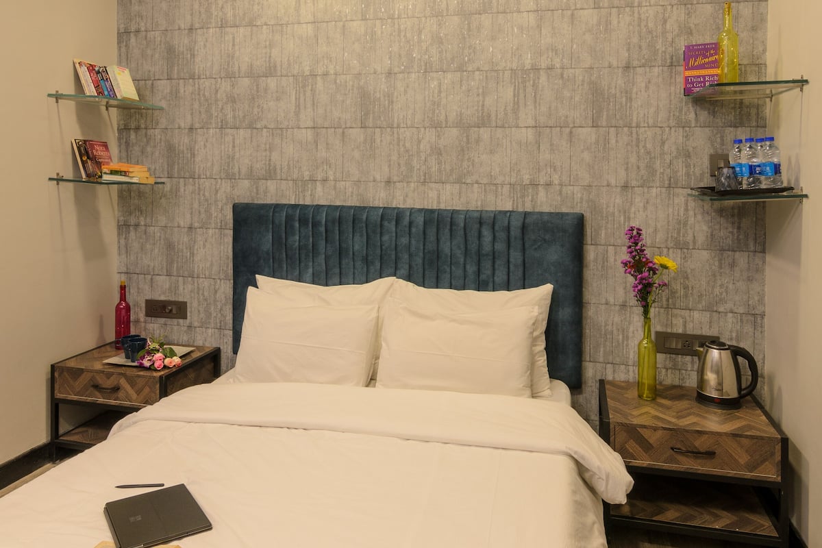 A neatly made bed with a blue headboard, a bedside table with a book and a bottle, and a wall-mounted shelf with books and a vase.