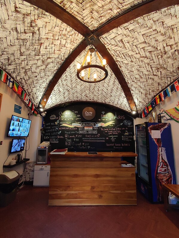 A hostel reception area with a check-in counter, a chandelier, a wall with hostel services information, and a television displaying a map. - Backpacker Cowies (Mumbai)