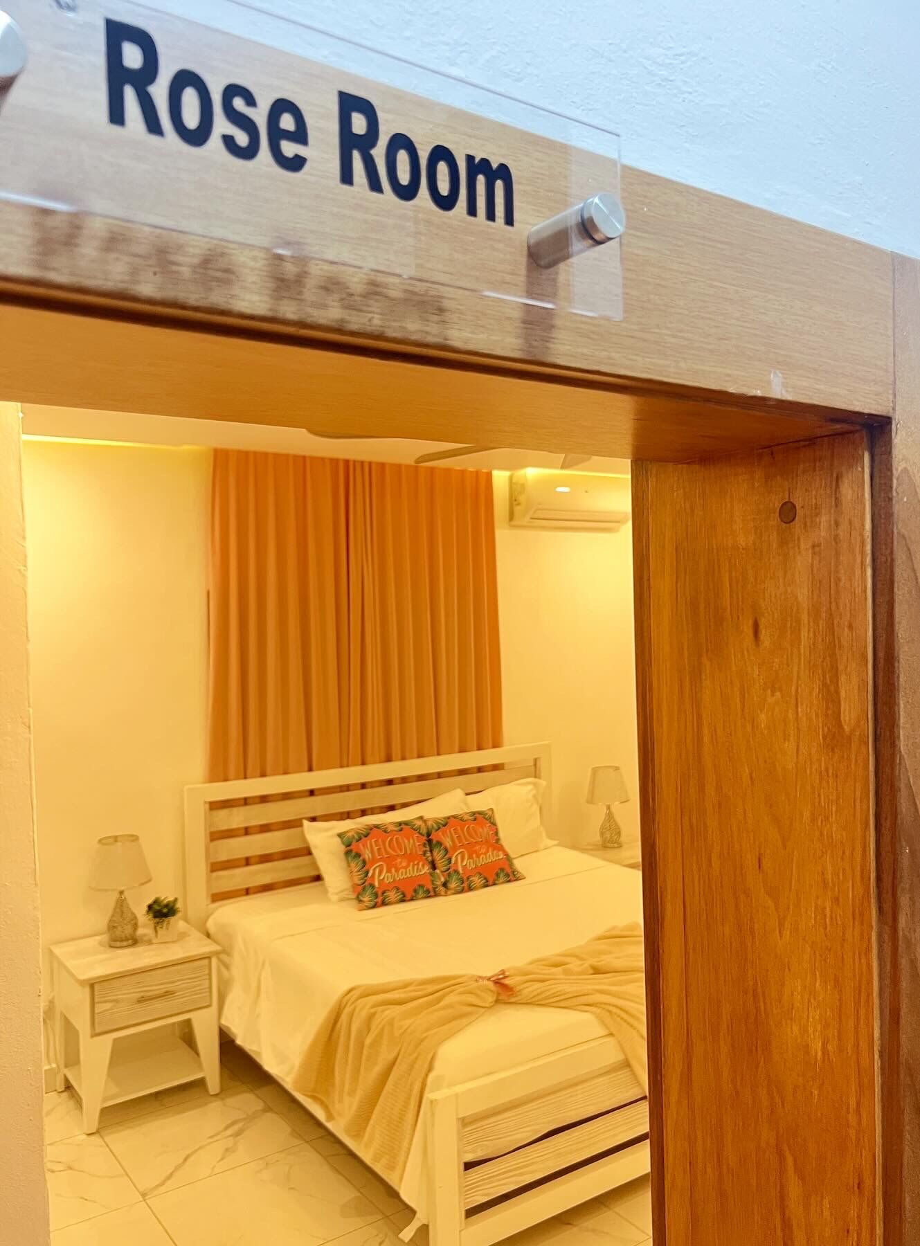 Ein Rosen-Zimmer mit einem Bett, zwei Nachttischen, einem Schreibtischchen, einem Fenster mit Vorhängen und einem Schild über der Tür.