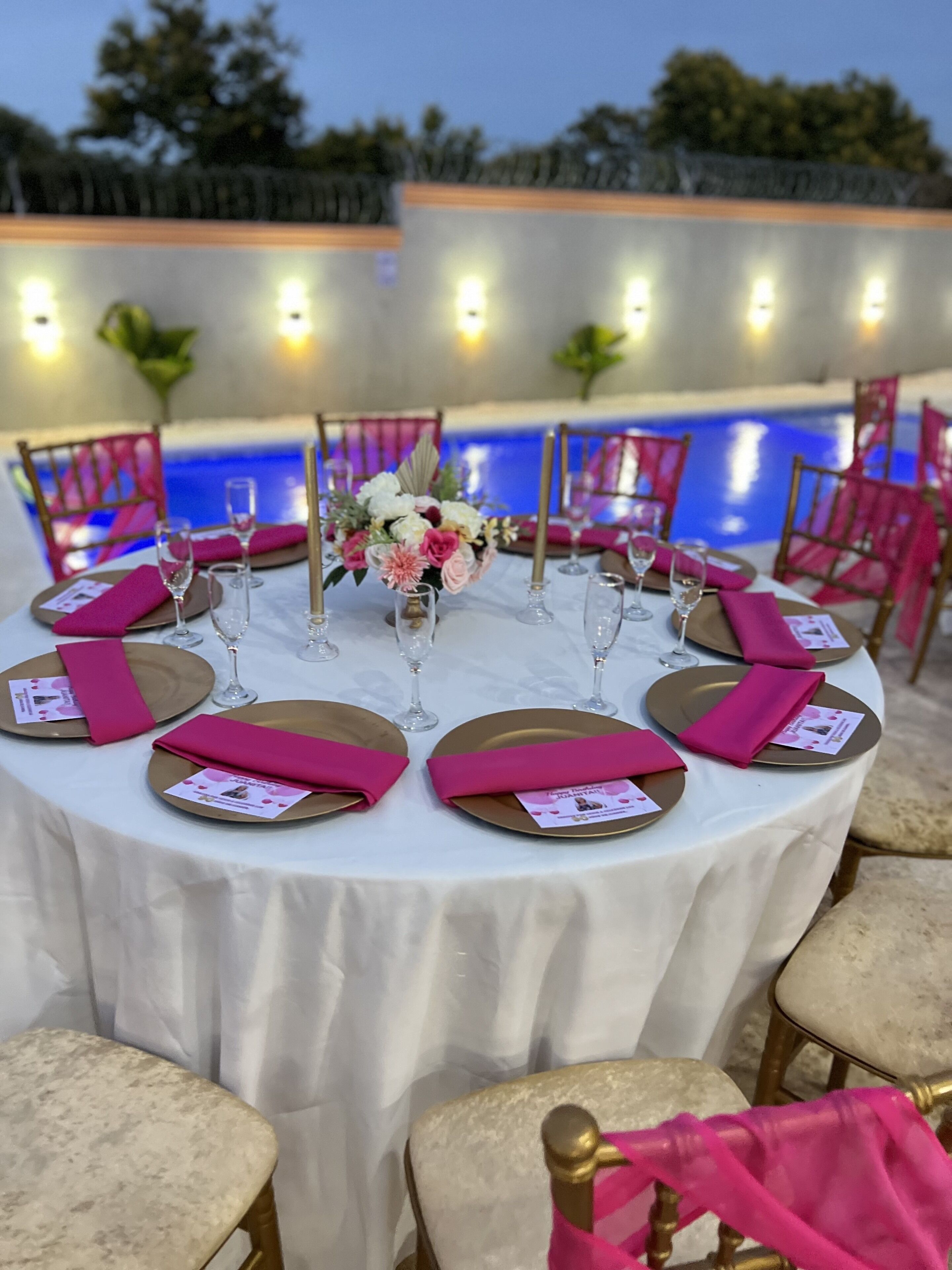 Een ronde eettafel gedekt voor een maaltijd, met roze servetten en gouden borden, omringd door stoelen en met een zwembad op de achtergrond.