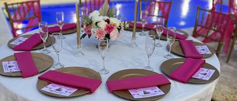 Een ronde eettafel gedekt voor een maaltijd, met roze servetten en gouden borden, omringd door stoelen en met een zwembad op de achtergrond.