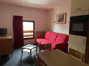 A compact hotel room with a red sofa, a small wooden table, a TV on a desk, and a kitchenette with an oven.