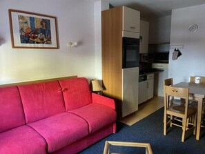 A compact living space with a red sofa, a small dining table with chairs, a kitchenette, and a framed picture on the wall.