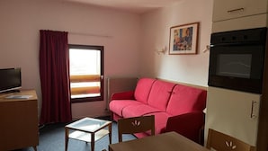 A compact hotel room with a red sofa, a small wooden table, a TV on a desk, and a kitchenette with an oven. - 150m from ski slopes, 2nd floor, view resort, fitness, swimming pool, sauna, tv, ski locker, 29m² (Eaux-Bonnes)