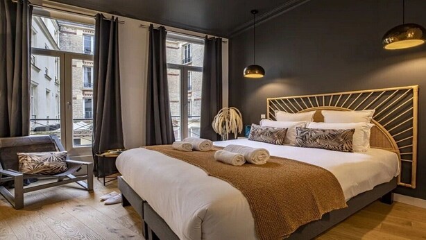 A modern bedroom with a large bed, a chair, and a view of the city through the window. - Marais Rue de Turenne  and its very particular charming atmosphere.
 (Paris)
