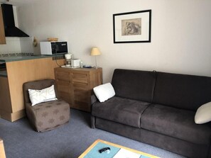 Un espacio habitable compacto con un sofá, un sillón pequeño, una kitchenette y un cuadro en la pared.