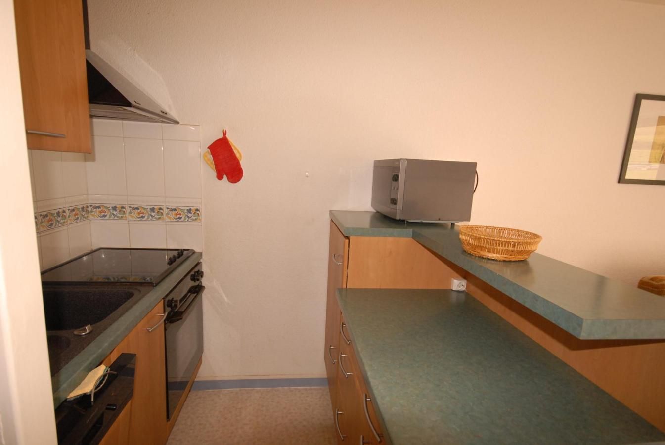 Een kleine keuken met een gasfornuis, oven en magnetron. Een rode ovenhandschoen hangt aan de muur, en er staat een geweven mand op het aanrecht.