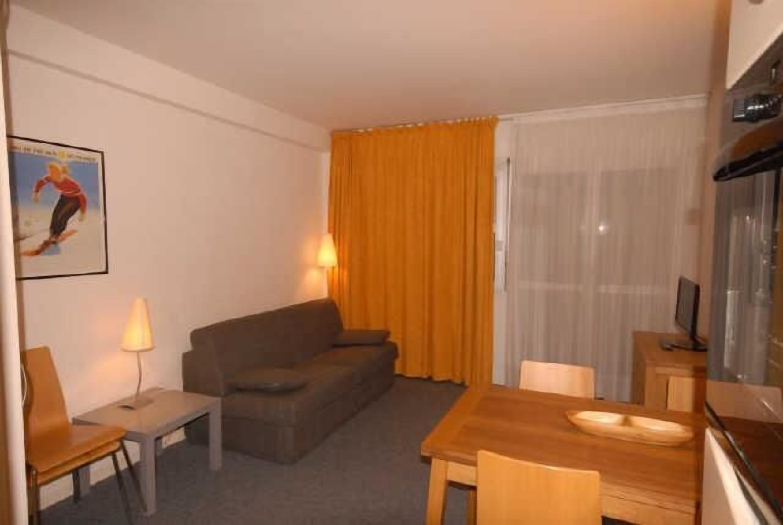 A hotel room with a sofa, a small table with a lamp, a wooden dining table, and a TV.