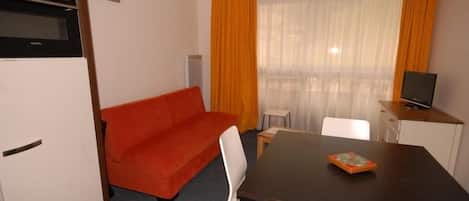 Ein gemütlicher Raum mit Sofa, Esstisch, Fernseher und einem Fenster mit Vorhängen.