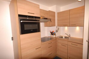 Uma cozinha compacta com armários de madeira, forno embutido, pia e bancada com uma cesta de produtos de limpeza.