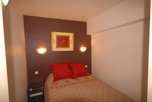 A small, neatly arranged bedroom with a bed, two pillows, a wall-mounted light, and a framed picture.