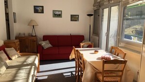 Un salon chaleureux avec un canapé rouge, une table à manger entourée de chaises et un meuble en bois. Les murs sont ornés de tableaux encadrés et une fenêtre offre une vue agréable.
