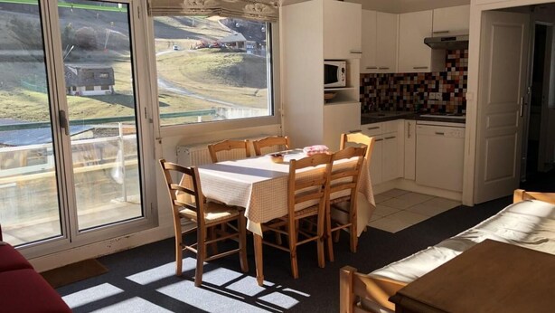Un espace repas avec une table et des chaises, une cuisine dotée de meubles blancs et d’un pare-feu en carrelage, et une vue dégagée grâce à de grandes fenêtres.