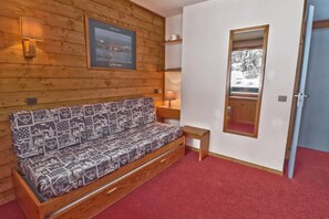 Quarto de cabana de madeira com sofá-cama, uma pequena mesa, abajur e um quadro com vista para uma cidade costeira.