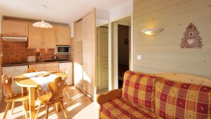 Un salon chaleureux avec un canapé en bois et une table à manger avec des chaises. On aperçoit, au fond, une cuisine avec des meubles en bois et un four à micro-ondes.