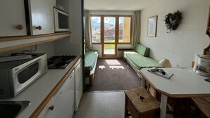 Eine kompakte Küche mit Mikrowelle, Herd und Spüle. Ein kleiner Esstisch mit Stuhl und eine Couch mit Blick ins Freie.