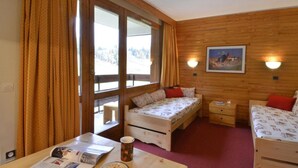 Una habitación acogedora con paredes de madera, una cama con ropa de cama floral, una mesita con un periódico y una taza, y un ventanal con cortinas.